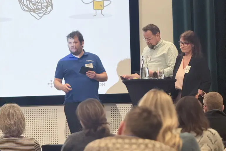 To menn og kvinne står på scenen foran publikum i konferansesalen.