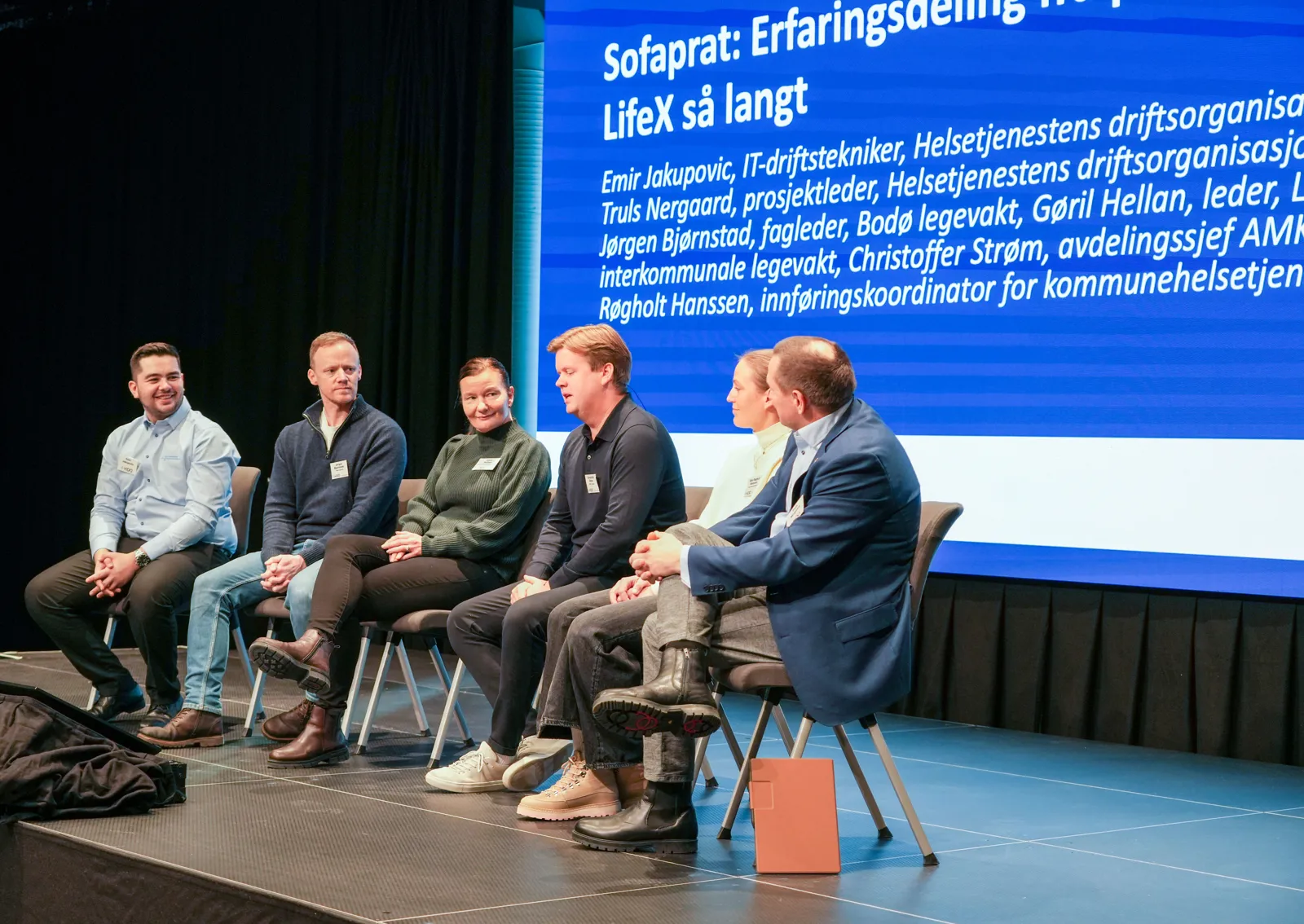 Panelsamtale på scene med sju deltakere foran en skjerm med informasjon om LifeX‑erfaringsdeling.