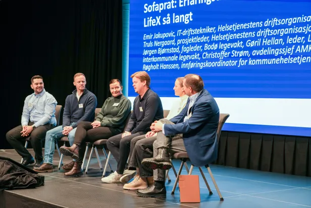 Panelsamtale på scene med sju deltakere foran en skjerm med informasjon om LifeX‑erfaringsdeling.