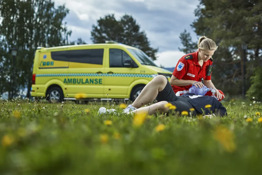 Ambulansepersonell som hjelper en person, ambulanse i bakgrunnen