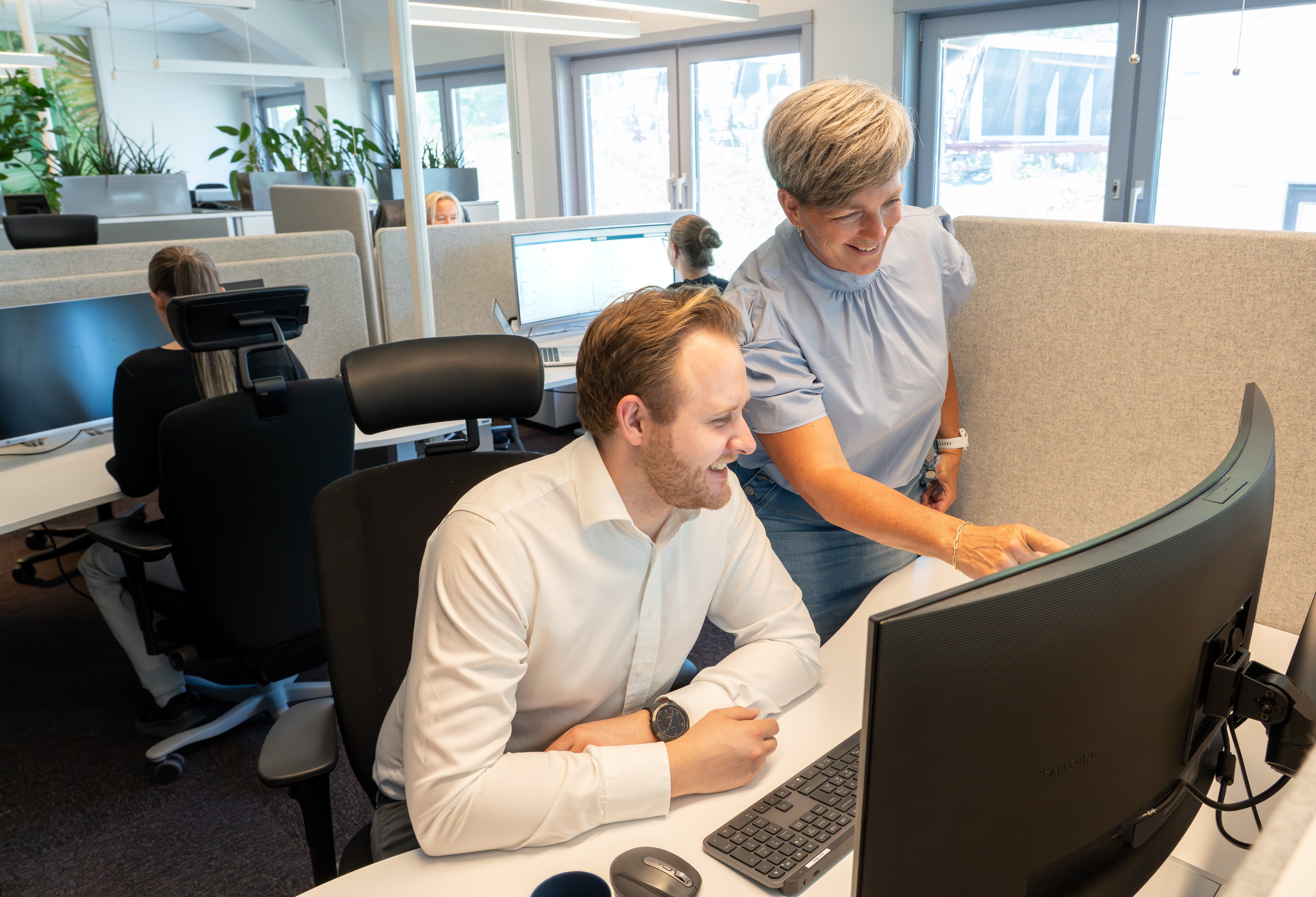 To kolleger samarbeider ved en kontorpult i et moderne, lyst kontorlandskap. En mann sitter foran en buet skjerm mens en kvinne står ved siden av og peker på skjermen. Flere ansatte jobber i bakgrunnen, og grønne planter er plassert rundt i rommet.