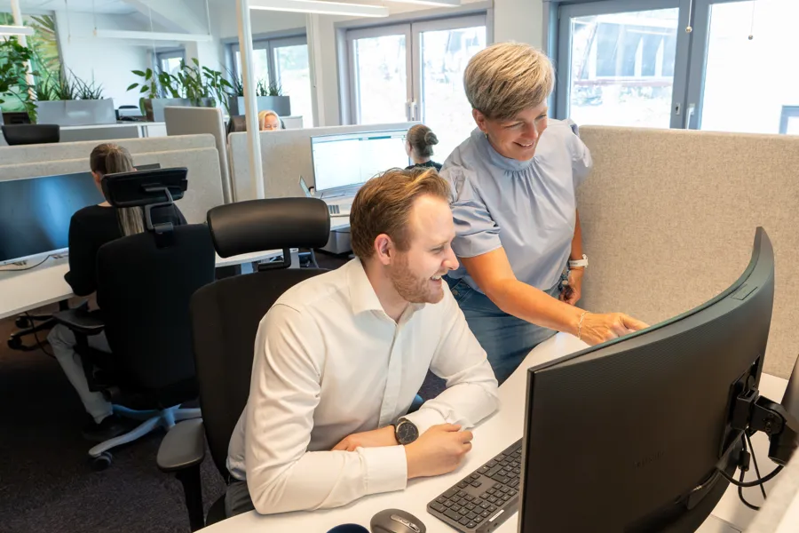 To kolleger samarbeider ved en kontorpult i et moderne, lyst kontorlandskap. En mann sitter foran en buet skjerm mens en kvinne står ved siden av og peker på skjermen. Flere ansatte jobber i bakgrunnen, og grønne planter er plassert rundt i rommet.