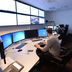 bilde av to mennesker som sitter på kontoret ved hver sin pult med mange skjermer