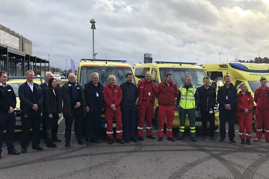 Bilde av en gruppe mennesker foran ambulanser. Noen i ambulanseklær og noen i dress
