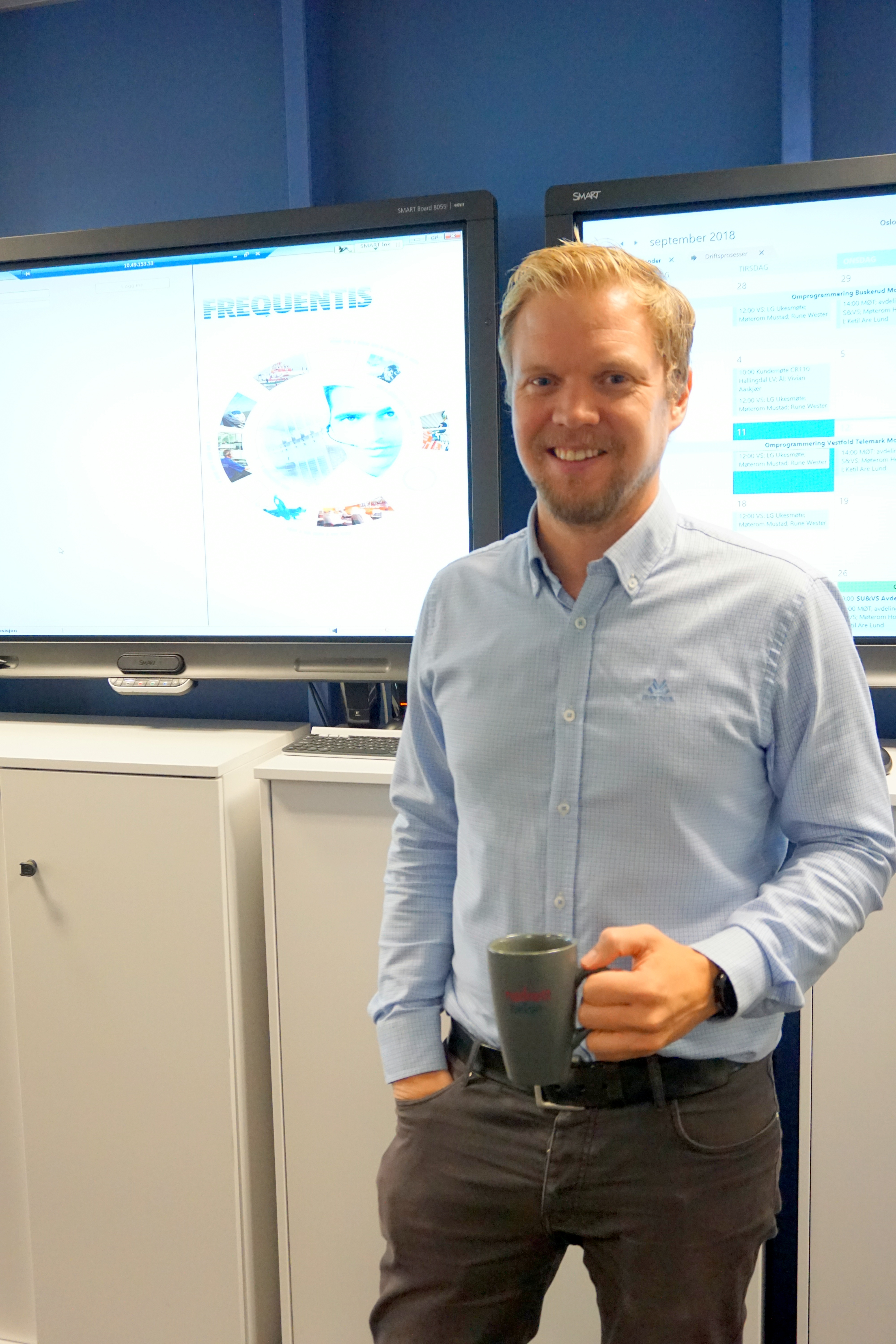 Person med kaffekopp står foran to skjermer i et møterom.