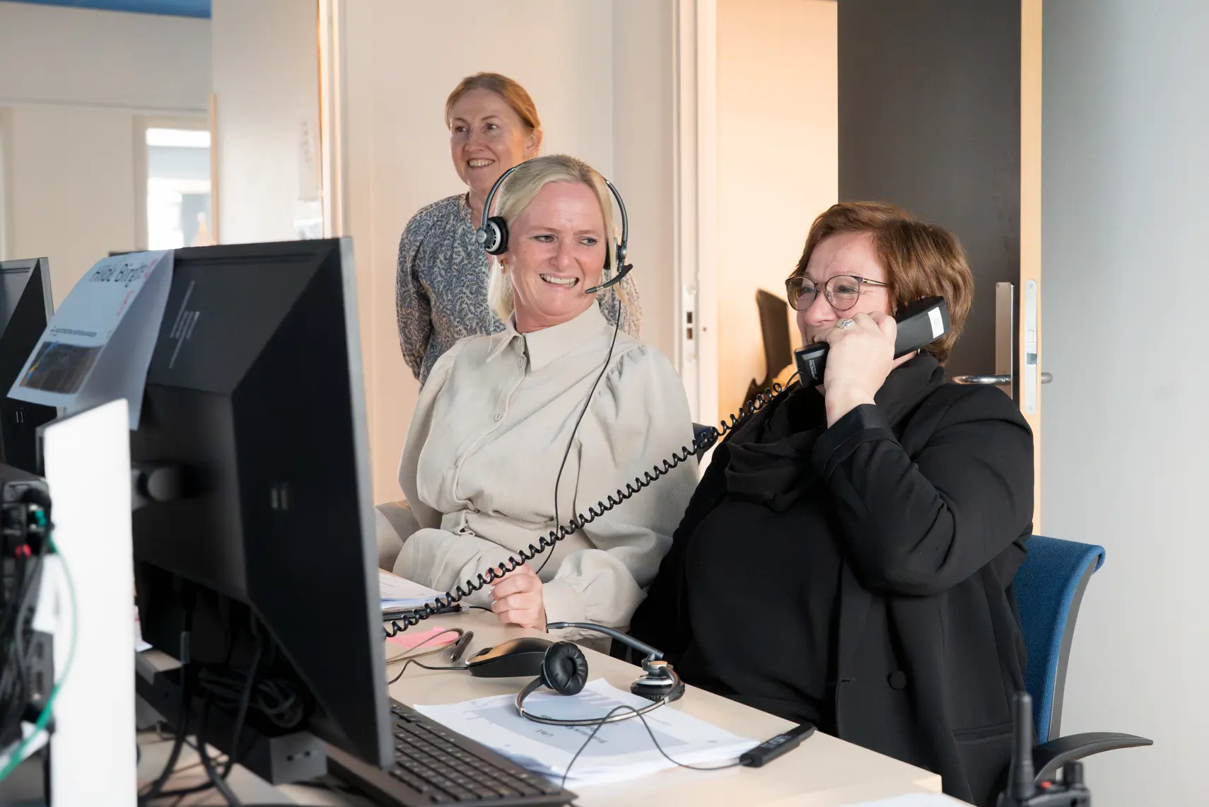 tre personer som sitter ved et skrivebord med dataskjermer og telefoner. To av personene har på seg hodetelefoner, og en av dem snakker i telefonen. Den tredje personen står bak de to andre og ser på skjermene.