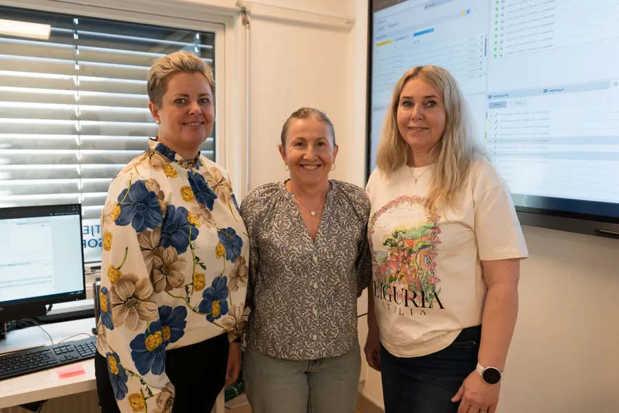 Tre damer står sammen i et rom med datamaskiner. To skjermer er synlige i bakgrunnen, en på venstre side og en stor skjerm på høyre side.