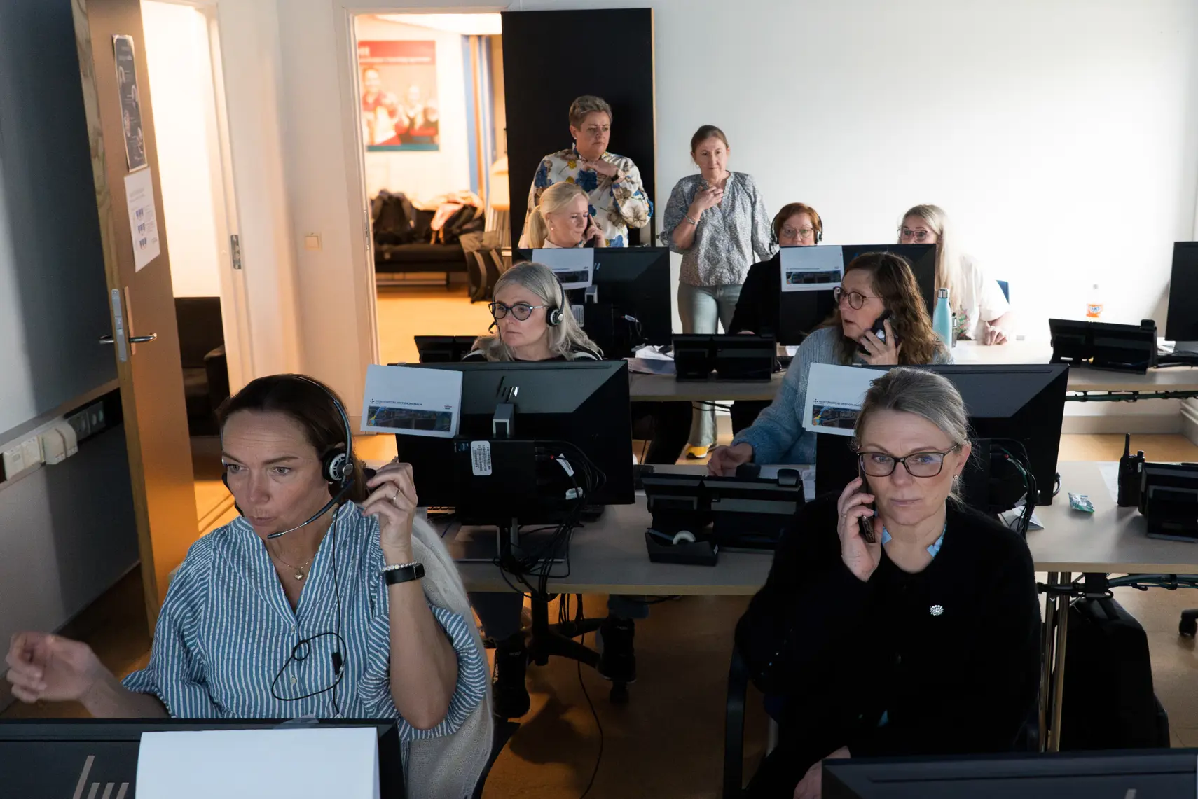 Et rom med flere personer som sitter ved datamaskiner. Noen av har på seg hodetelefoner og er engasjert i opplæring. Instruktører står helt i bakerst i lokalet.