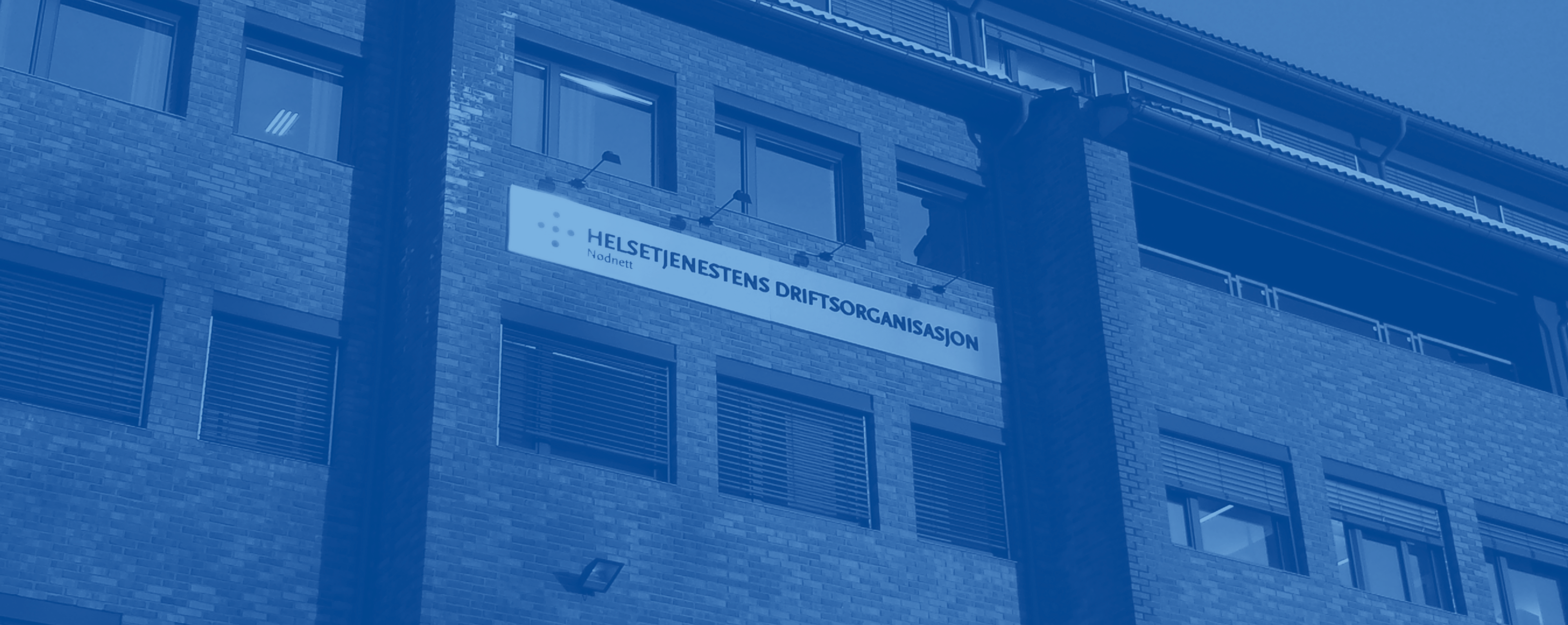 Fasaden til et murbygg med flere etasjer og store vinduer. Midt på bygget henger et skilt med teksten «Helsetjenestens driftsorganisasjon for nødnett». Bildet er tonet i blått og fotografert nedenfra.