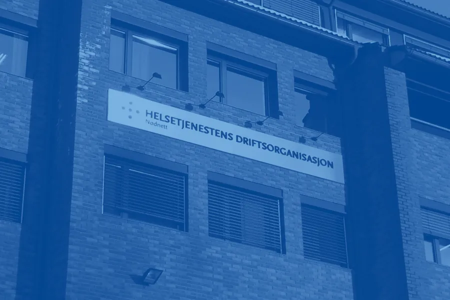 Fasaden til et murbygg med flere etasjer og store vinduer. Midt på bygget henger et skilt med teksten «Helsetjenestens driftsorganisasjon for nødnett». Bildet er tonet i blått og fotografert nedenfra.