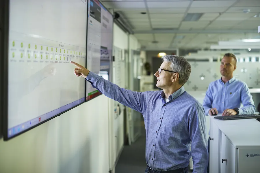 En mann peker på en stor skjerm med data i et moderne kontorlandskap, mens en kollega står i bakgrunnen ved en pult med tastatur.