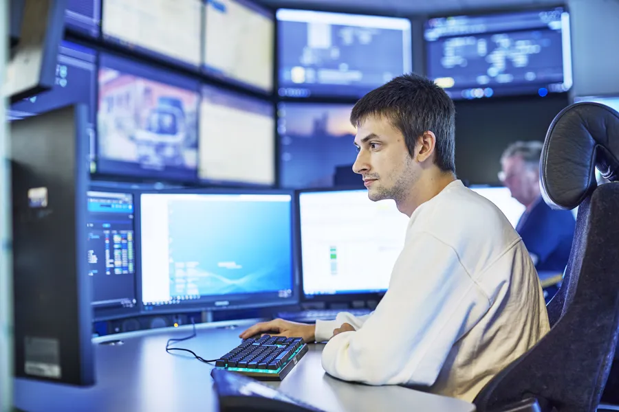 En person sitter foran flere dataskjermer i et kontrollrom. Personen har en hvit genser og bruker tastaturet. Flere skjermer viser ulike typer informasjon og overvåkingsbilder.