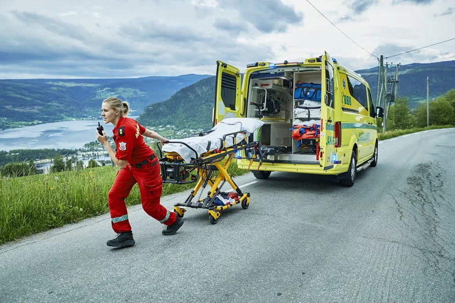 Nødsituasjon på en landevei: En ambulanse står parkert med åpne bakdører, og en båre trekkes bort av ambulansepersonell. Medisinsk utstyr er synlig inne i bilen. I bakgrunnen er fjell, innsjø og overskyet himmel.